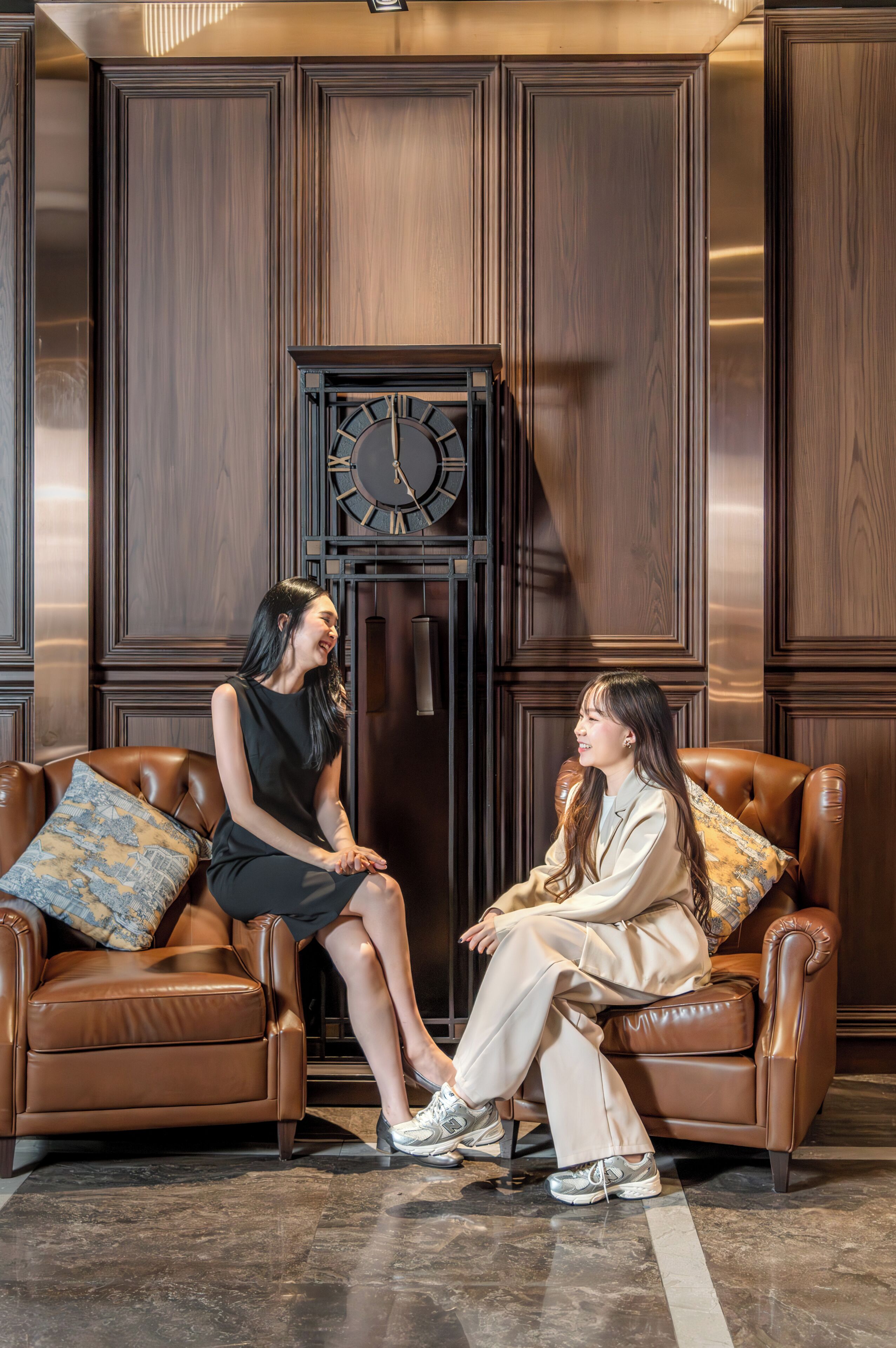 Two people seated on leather sofas in a room with wooden paneling and a large clock.