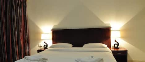 Quarto de hotel com uma cama grande, duas mesas de cabeceira com luminárias e uma cortina à esquerda.