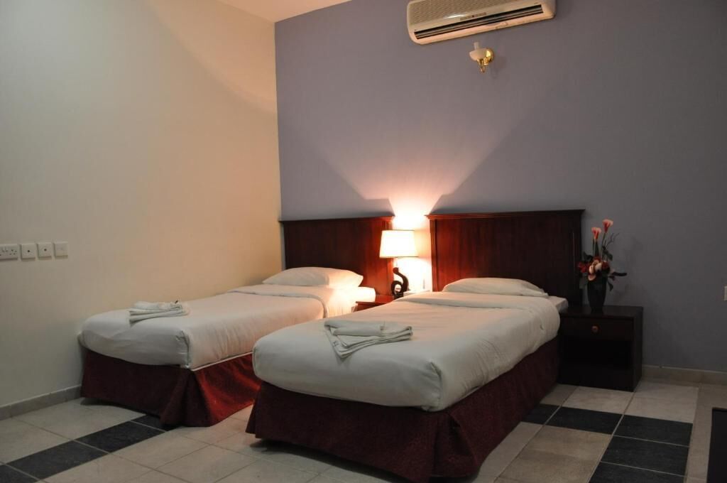 A hotel room with two single beds, a nightstand with a lamp, and an air conditioning unit on the wall.