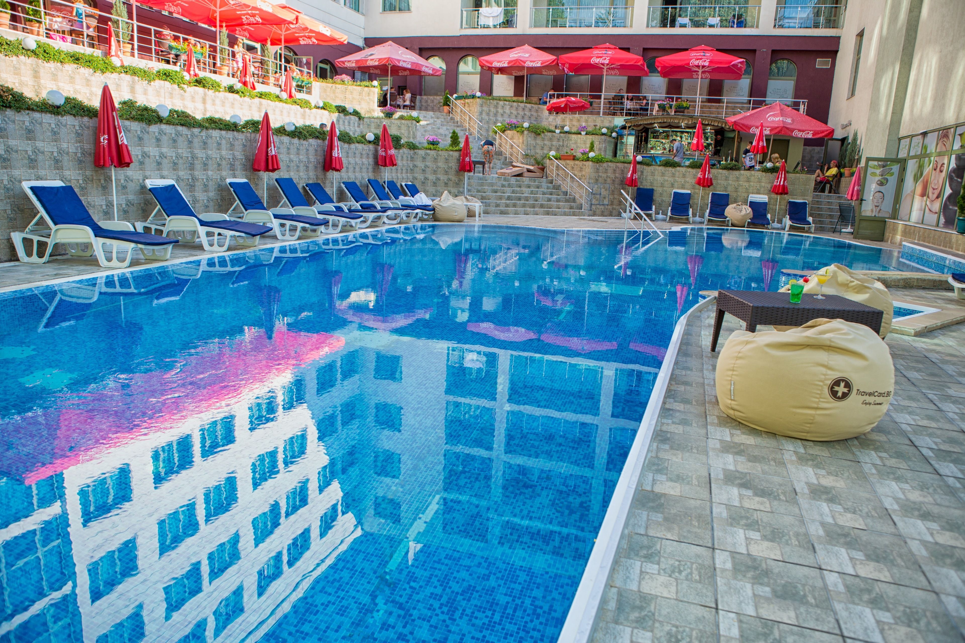 Una piscina con sillas de descanso y sombrillas, rodeada por un edificio de fachada de piedra y sombrillas rojas.