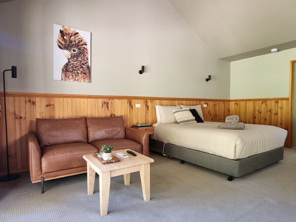 A room with a brown leather sofa, a wooden table with a plant, a bed with a grey mattress, and a wall-mounted picture.