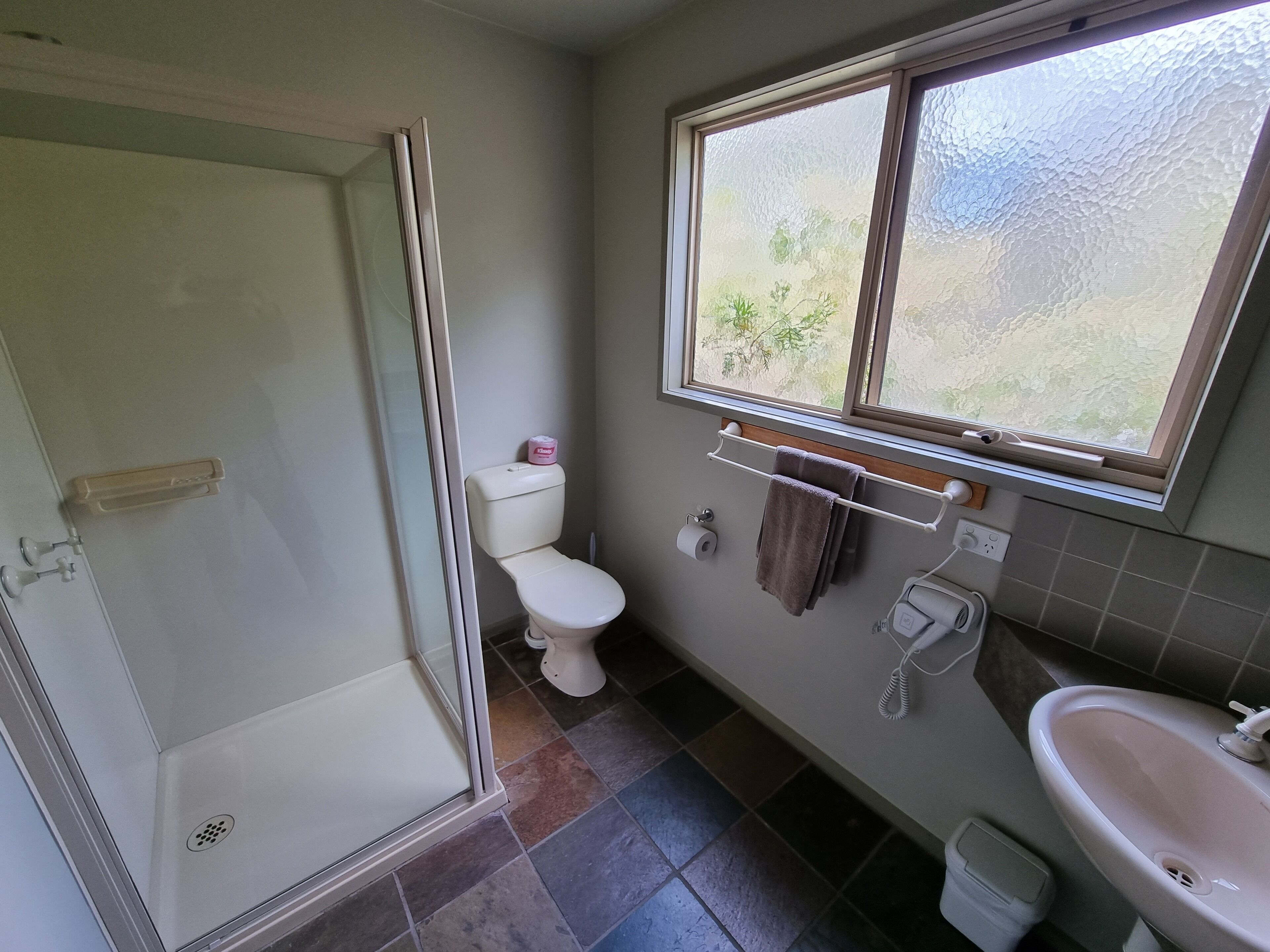 Een badkamer met een douchecabine van glas, een toilet, een wastafel en een raam met uitzicht op groen.
