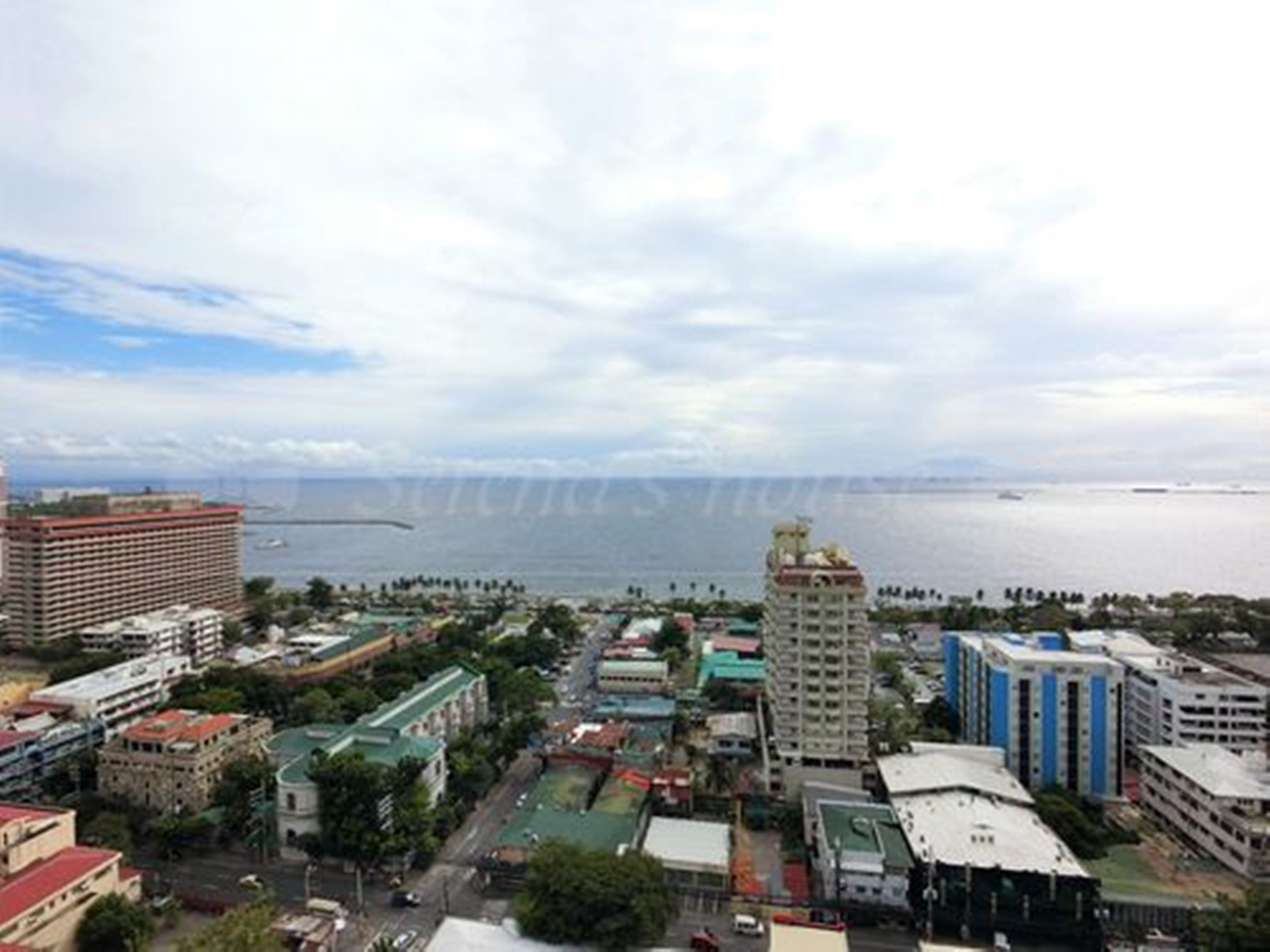 A coastal cityscape with a mix of modern and older buildings, a prominent bridge, and a large body of water in the background.