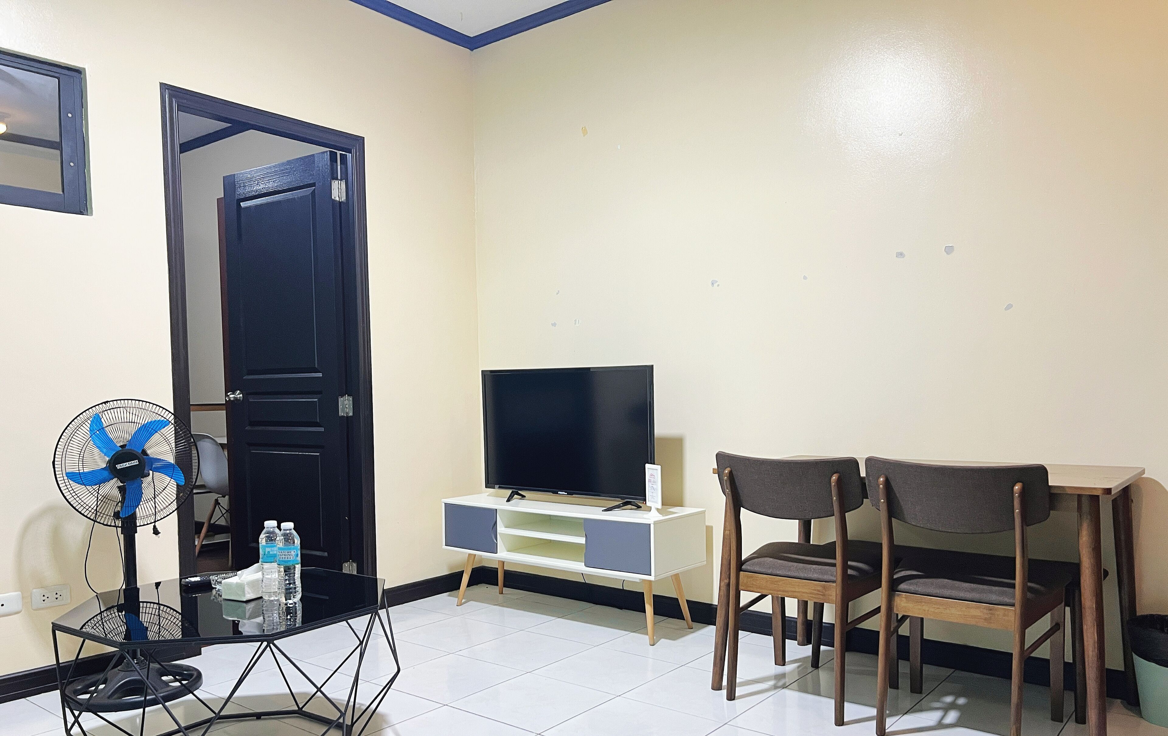 A room with a TV on a white stand, two wooden chairs, a glass table with a fan, and a door.