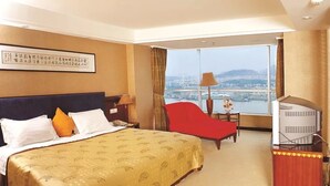A hotel room with a large bed, a red chair, a desk with a computer, and a view of a cityscape. - Shunde Grandview Hotel (Foshan)