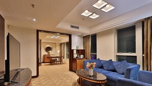 A hotel room with a blue sofa, a small round table with a vase of flowers, a TV, and a minibar. - Shunde Grandview Hotel (Foshan)