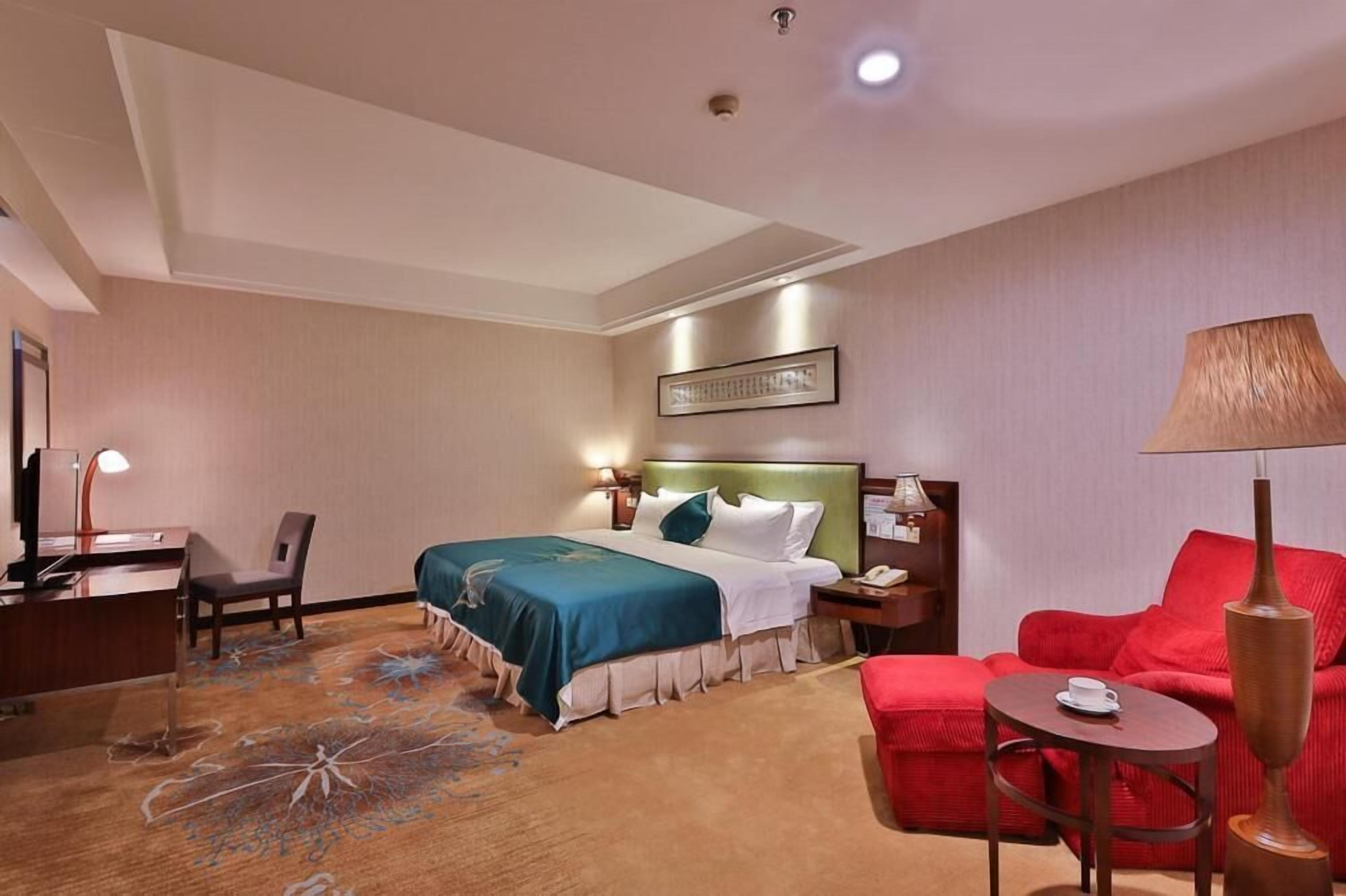 A hotel room with a bed, a desk, a chair, a red sofa, and a small table with a lamp.