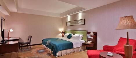 A hotel room with a bed, a desk, a chair, a red sofa, and a small table with a lamp.