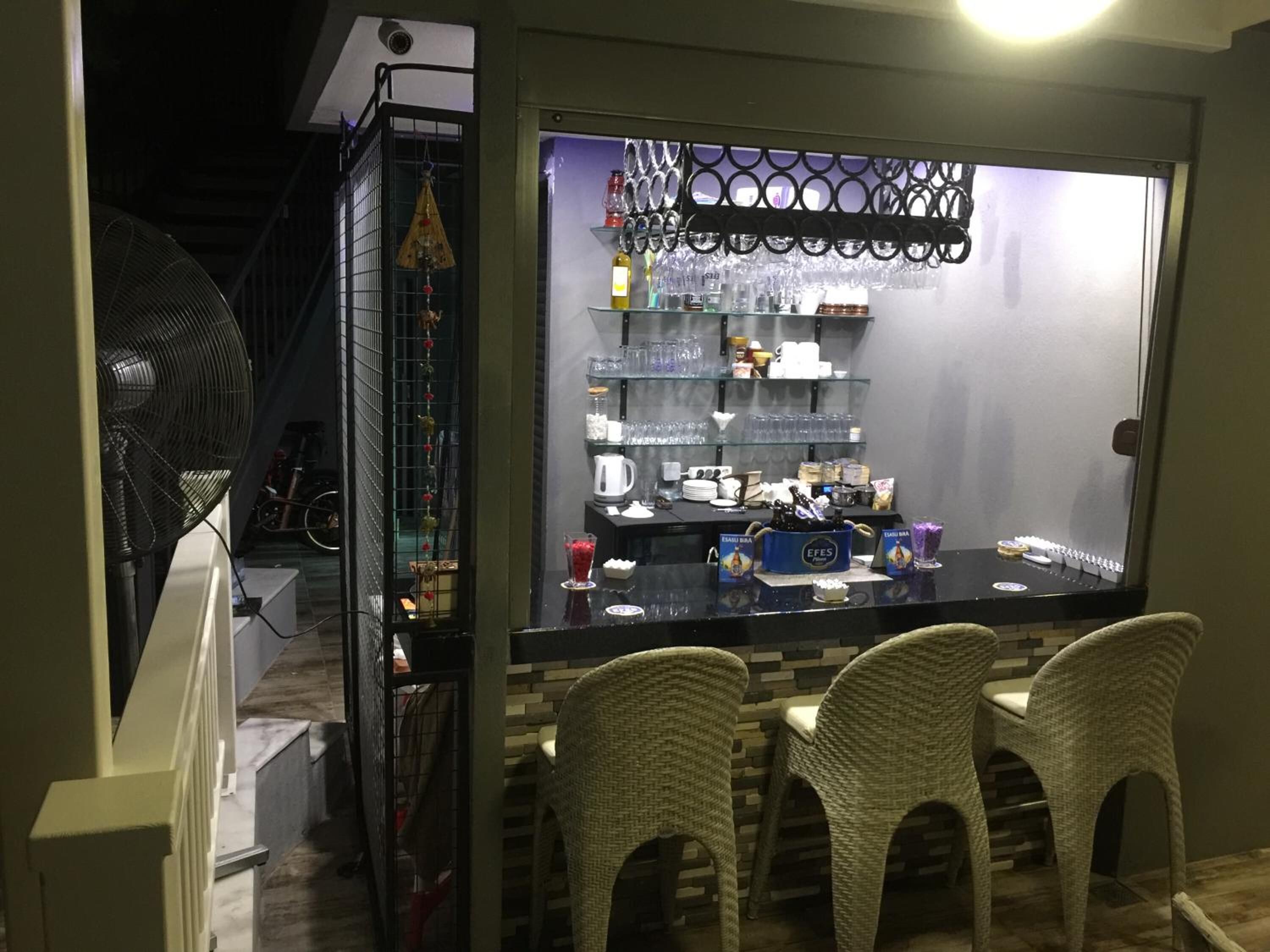 Un área de bar con sillas de mimbre, una barra con diversas botellas y vasos, y una estantería metálica decorativa arriba.