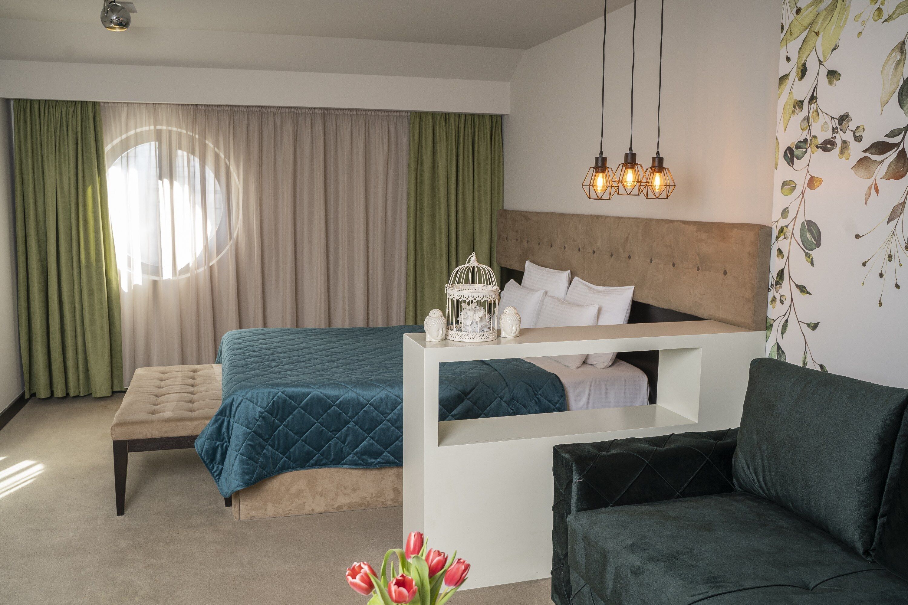 Ein Schlafzimmer mit Bett, Sitzbank und Sofa. Ein Fenster mit Vorhängen, eine Wand mit Blättermuster und eine Deckenleuchte.