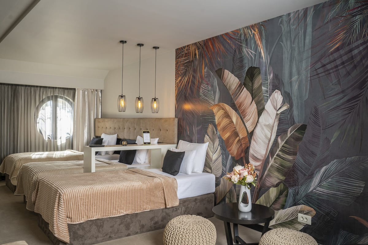 A hotel room with two beds, a large tropical leaf mural, a round window, and a small table with a vase.