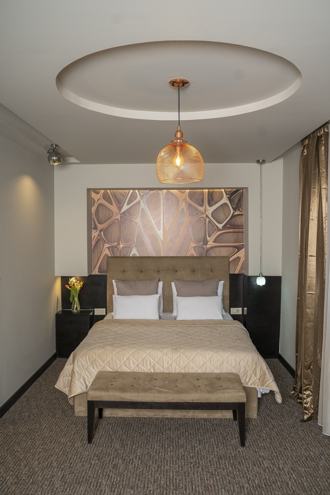 Habitación de hotel con una cama grande, cabecera de madera, lámpara de techo redonda y pared con diseño abstracto.