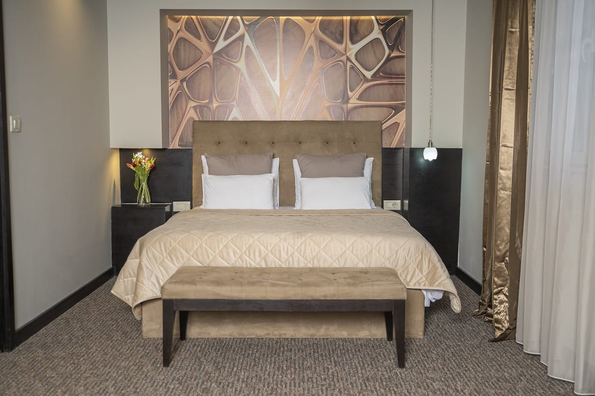 A hotel room with a large bed, a bench, a nightstand with a vase of flowers, and a decorative wall panel.