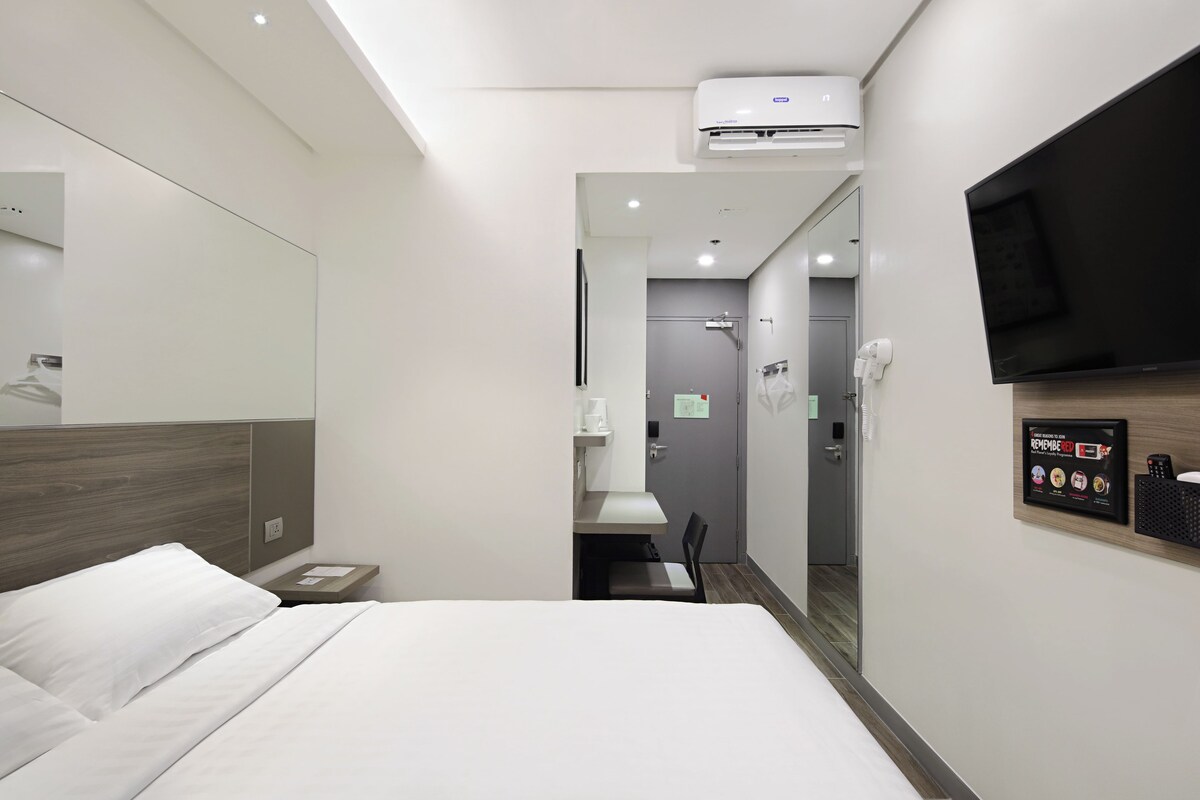 A modern hotel room with a large bed, a flat-screen TV mounted on the wall, a small bedside table, and an air conditioning unit.