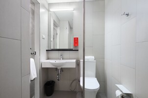 Un baño moderno con lavamanos, espejo y retrete blancos. Se observa un cubo de basura y un perchero para toallas.