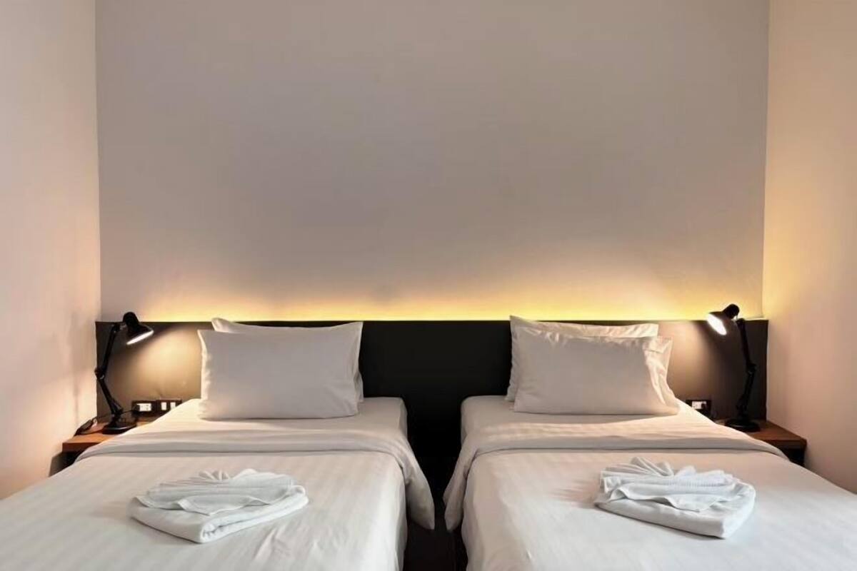 A hotel room with two single beds, each with a pillow and a folded towel. There are bedside lamps on either side of the beds.
