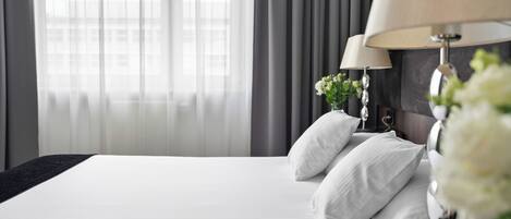 Ein Hotelzimmer mit einem großen Bett, zwei Nachttischen mit Lampen und einem Fenster mit durchsichtigen Vorhängen.