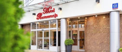 A hotel entrance with a glass canopy, a large potted plant, and a sign indicating room number 131.