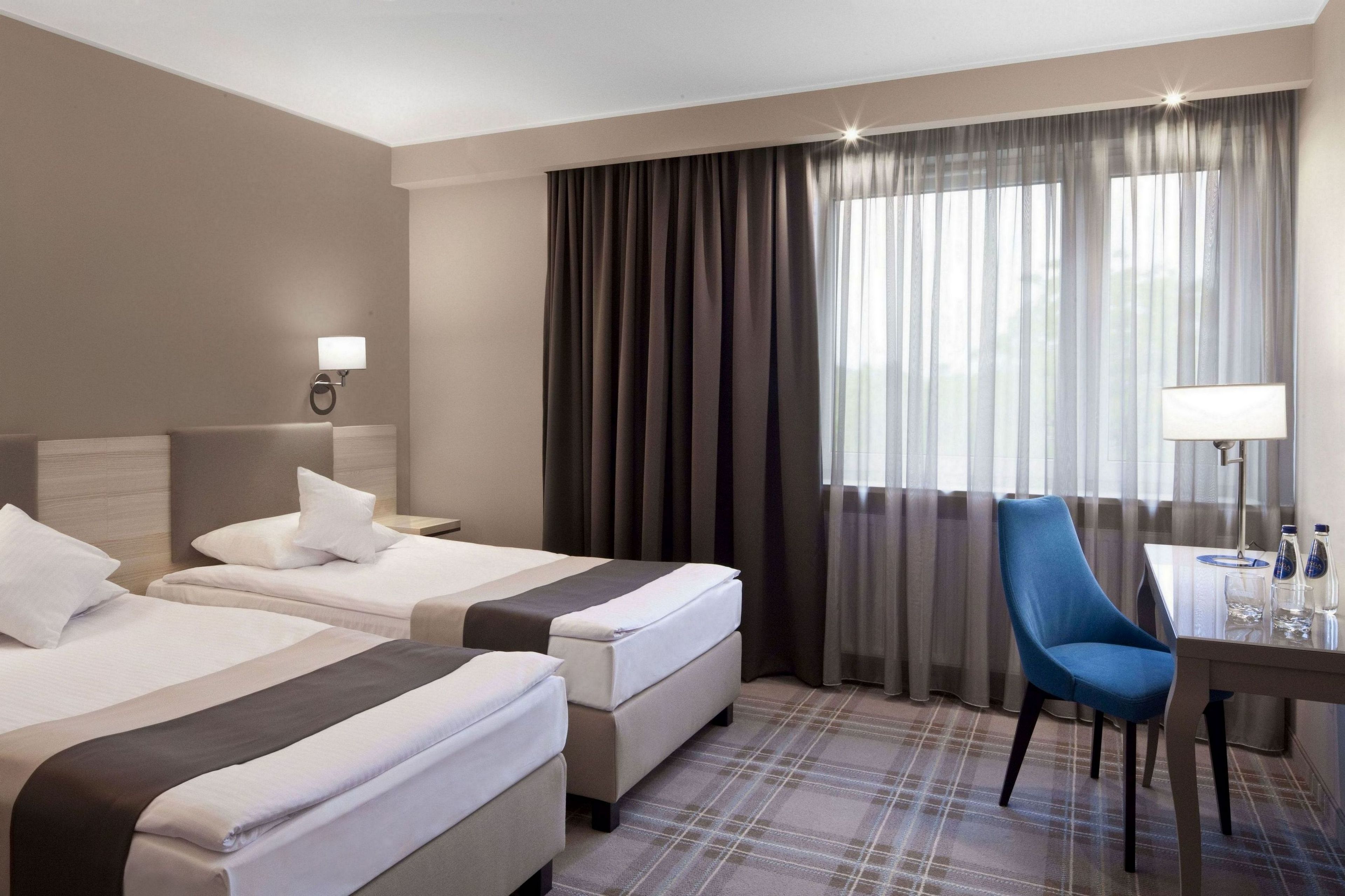 A hotel room with two beds, a desk, a chair, and a large window with curtains.