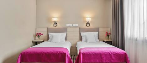 A hotel room with two beds, each with a pink bedspread, a headboard, bedside tables with lamps, and a checkered floor.