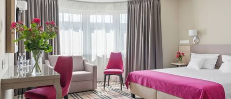 A hotel room with a bed, two chairs, a desk, a lamp, and a vase with flowers.