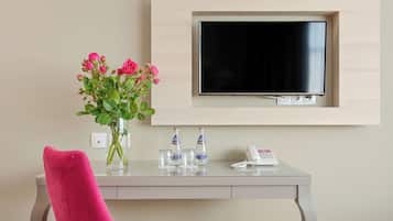 Habitación de hotel con escritorio, una silla rosa, un jarrón con flores, un televisor de pantalla plana montado en la pared y un teléfono.