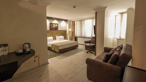 Quarto de hotel com uma cama, um sofá, uma escrivaninha com chaleira e um quadro na parede.