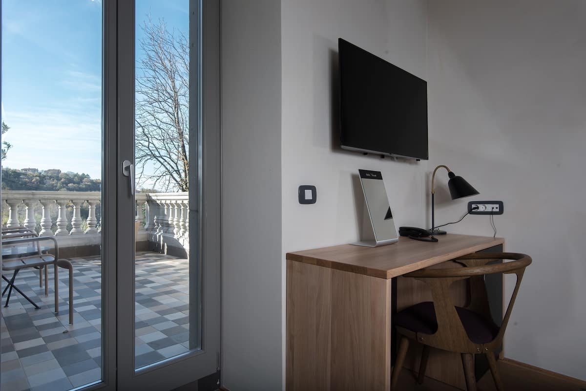Ein Zimmer mit Schreibtisch, Stuhl, Fernseher, Lampe und Blick auf einen Balkon mit Stühlen und Geländer.