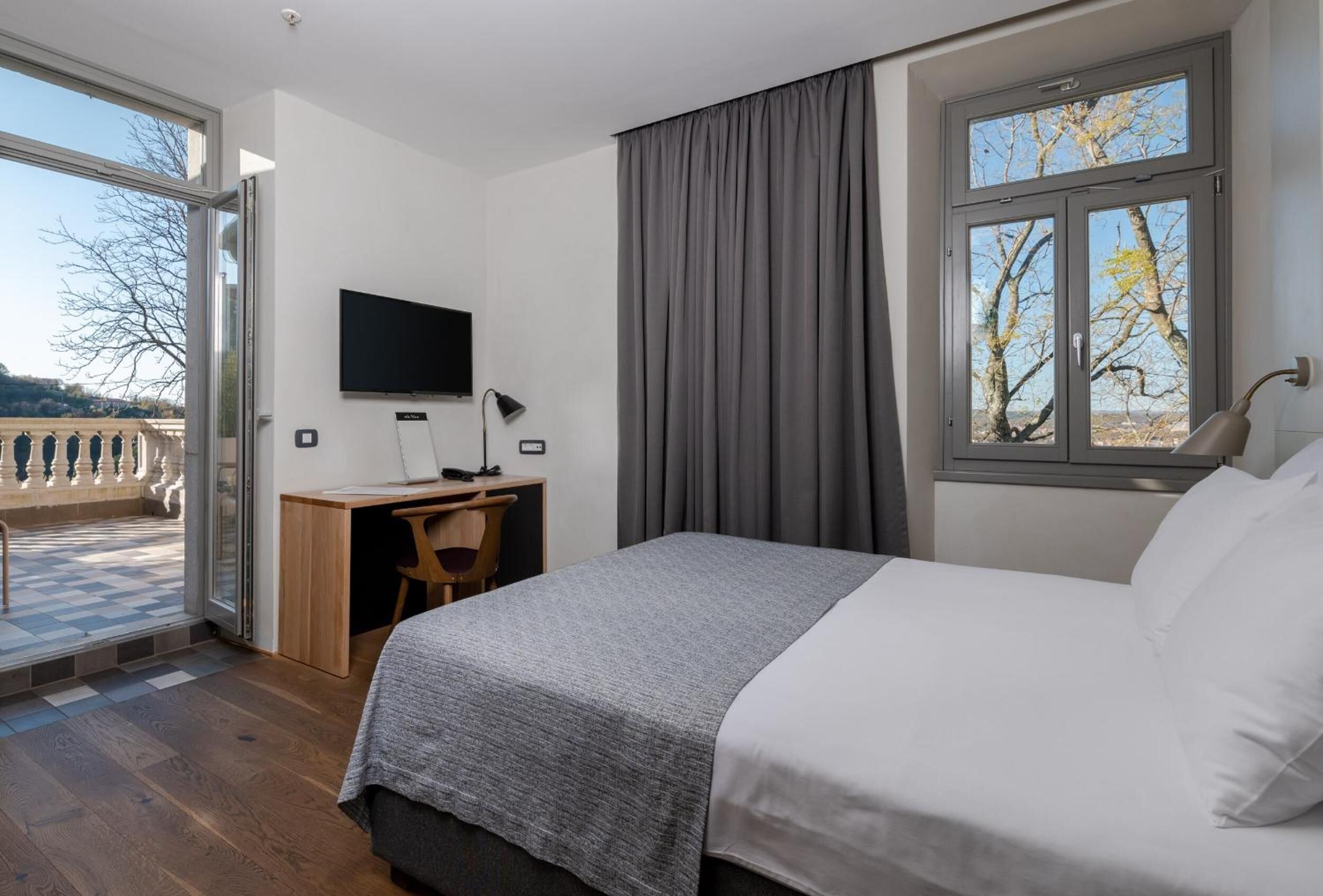 Una habitación de hotel con cama, escritorio, computadora, televisor y ventana con cortinas.