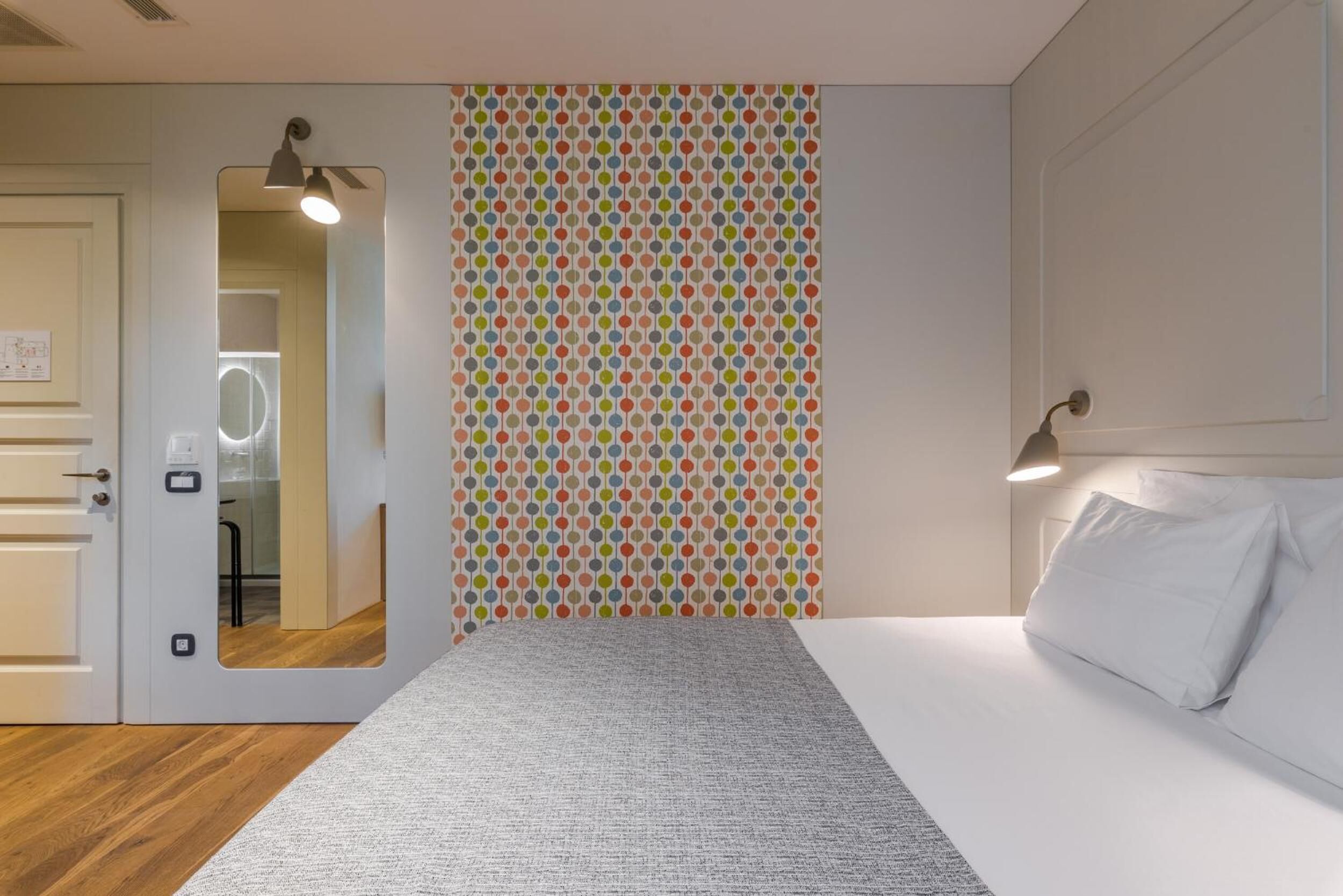 A hotel room with a bed, a mirror, a door, and a wall with a polka dot pattern.