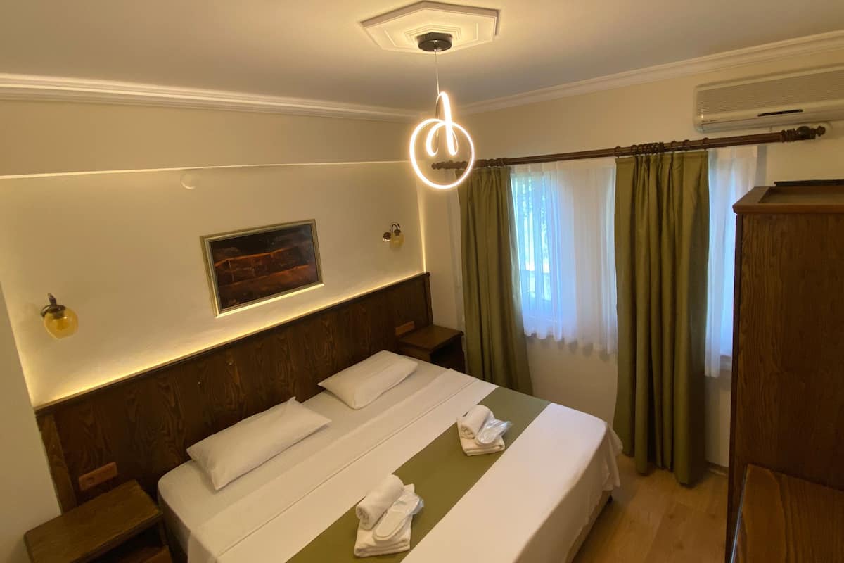 A hotel room with a neatly made bed, a headboard with built-in lighting, a painting on the wall, a wooden nightstand, and a large window with curtains.