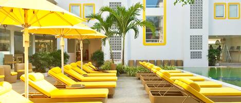 Zona de piscina con tumbonas amarillas y sombrillas, rodeada de plantas en macetas y un edificio con ventanas decorativas.