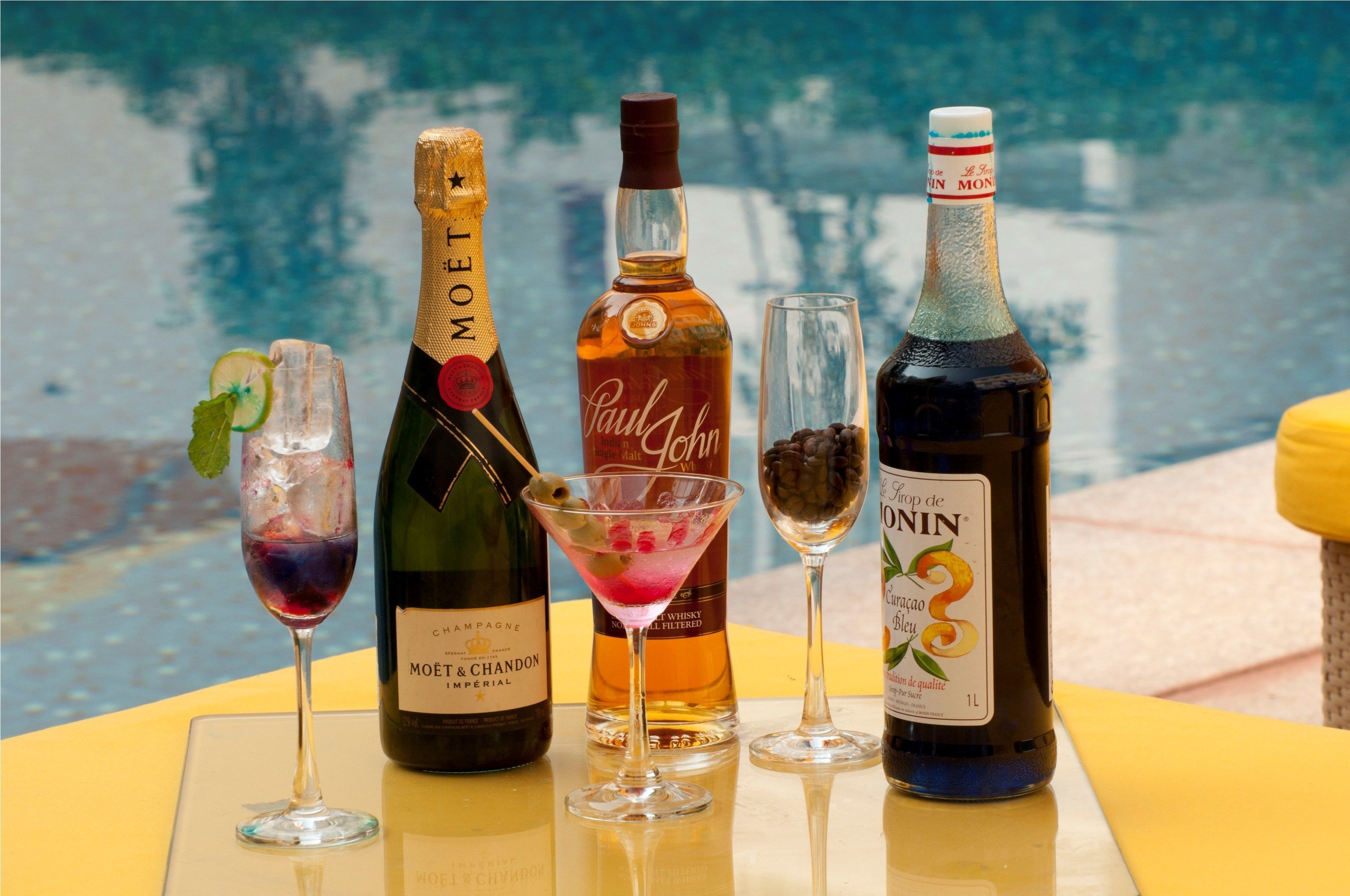 Une table avec cinq bouteilles d’alcool et deux verres de cocktails, placée au bord d’une piscine.