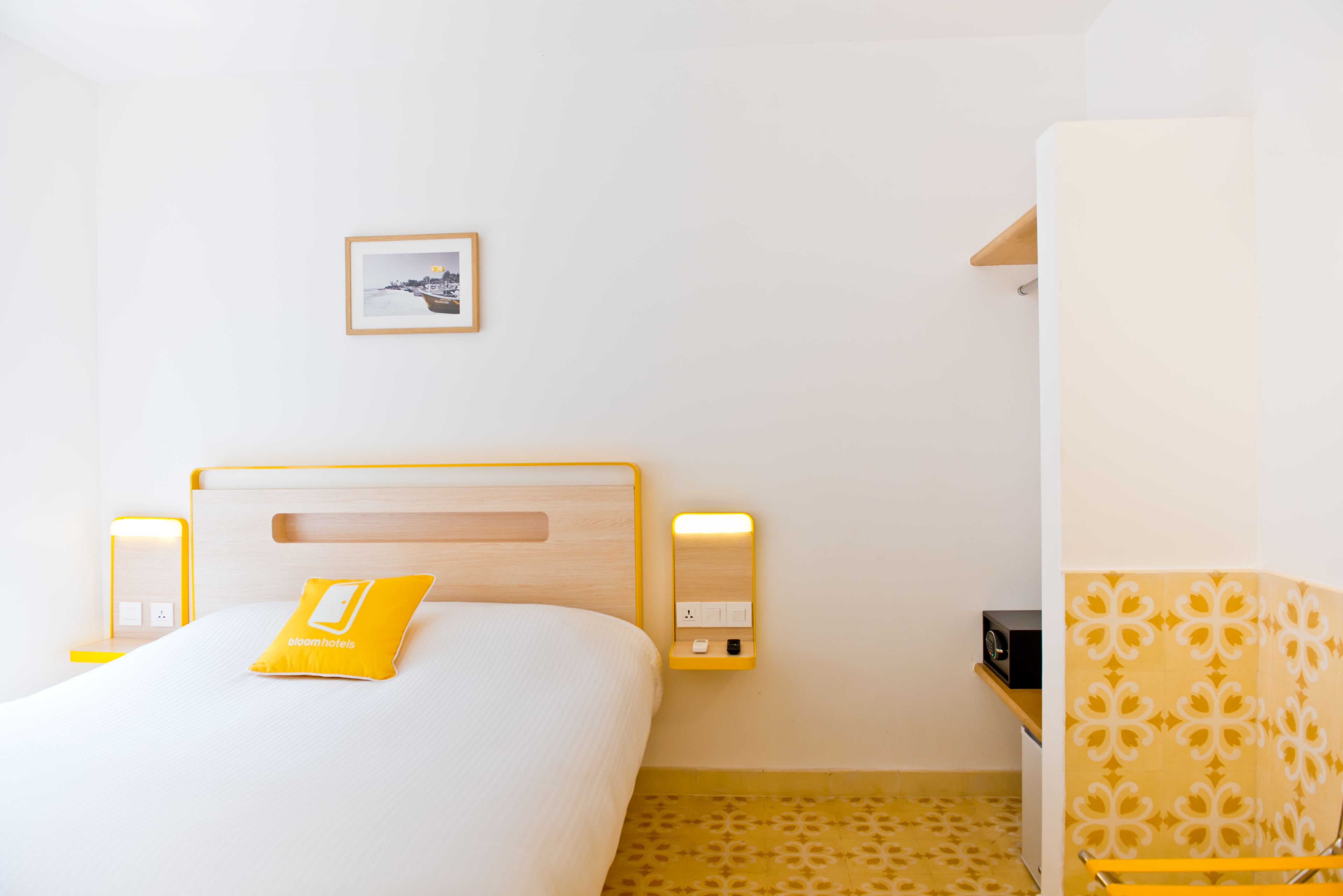 A hotel room with a bed, bedside lamps, a small table, a wall-mounted picture, and a patterned chair.
