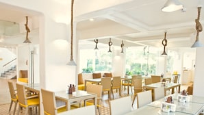 A modern dining area with glass tables and chairs, white walls, and hanging rope lights. - Bloom Hotel – Calangute (Calangute)