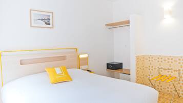 Habitación de hotel con cama, mesita de noche con lámpara, estante pequeño, cuadro en la pared y perchero.