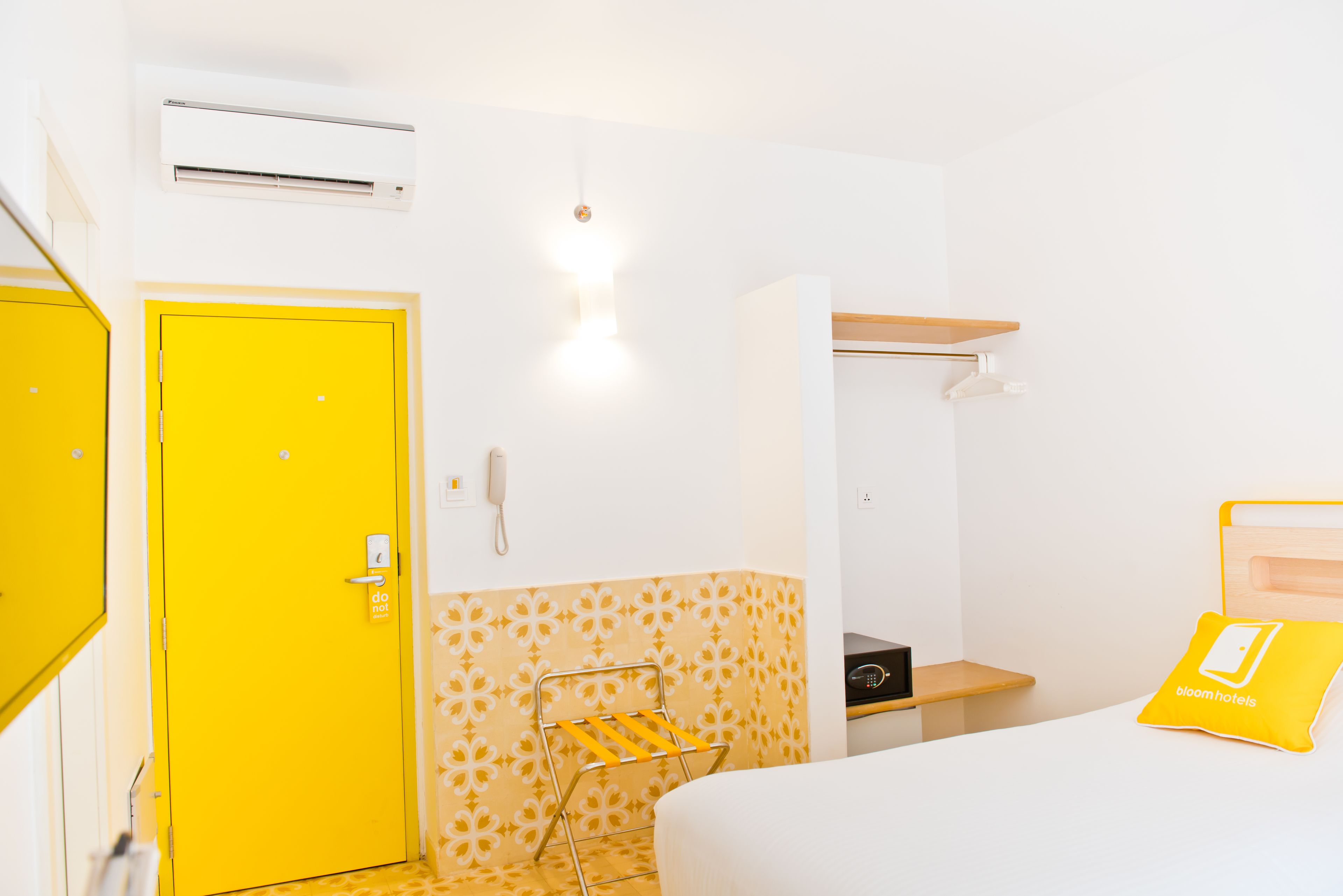 A hotel room with a yellow door, a bedside table, a bed, a wall-mounted air conditioner, a telephone, and a bedside lamp.