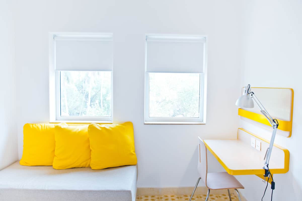 A room with a white sofa with yellow cushions, a desk with a yellow lamp, and two windows with white blinds.
