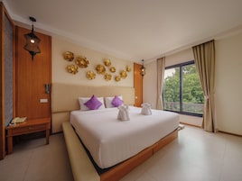 A hotel room with a large bed, a wooden bench, a telephone, and a view of greenery through the window.