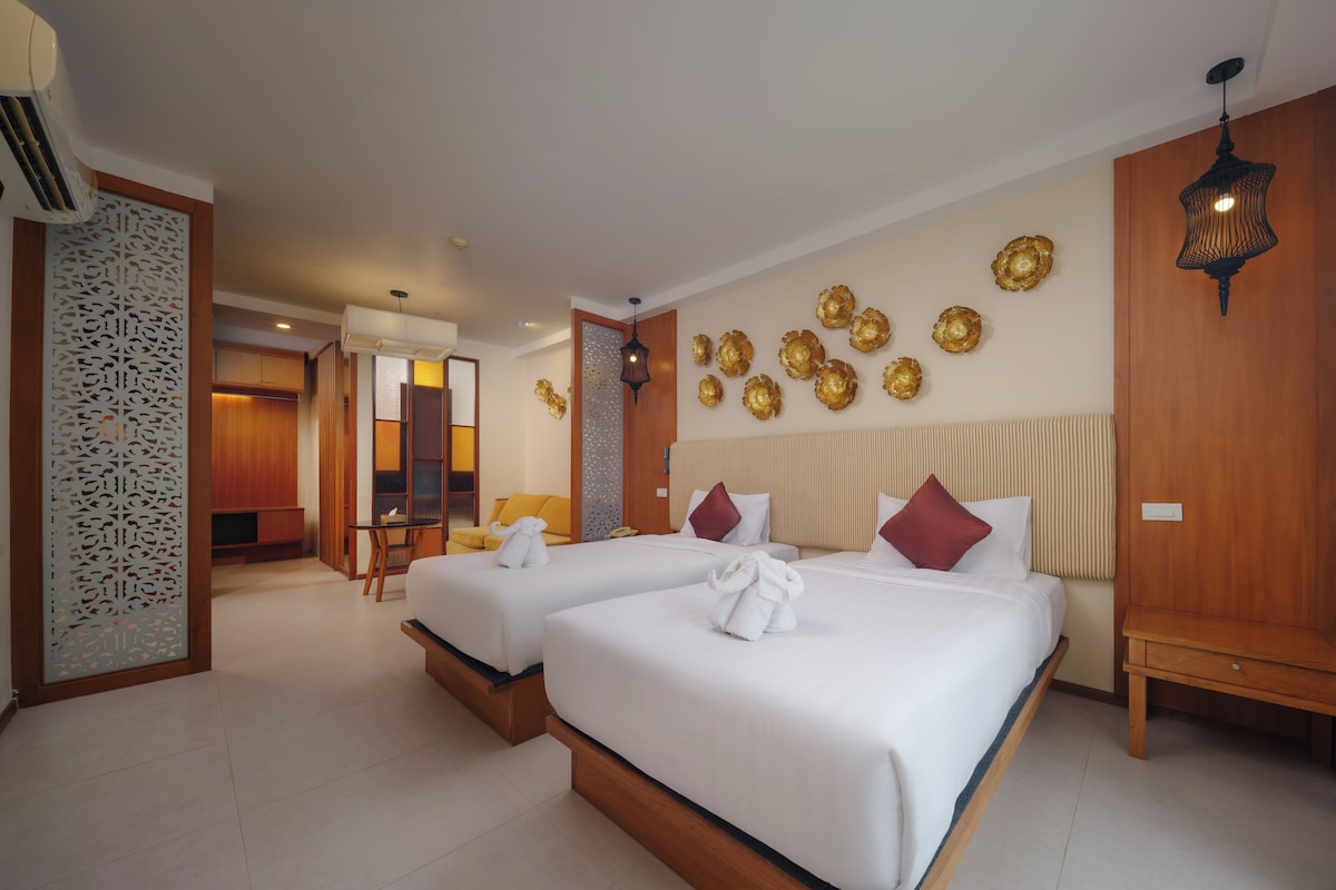 A hotel room with two beds, a wooden headboard, a decorative wall with golden ornaments, and a separate seating area with a sofa and table.