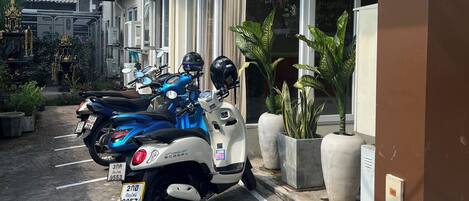 Un espacio de estacionamiento con scooters, la entrada de un edificio y plantas en macetas.