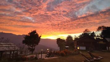 A sunset view with a mountain in the background, a wooden house, and a patio with chairs.