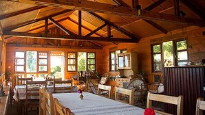 Interior rústico de una cabaña con vigas de madera, una mesa de comedor, sillas y una zona de cocina.