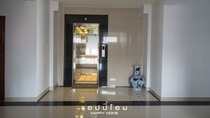 Um elevador moderno com visor digital, tapete de boas-vindas e um vaso decorativo num corredor limpo e bem iluminado.