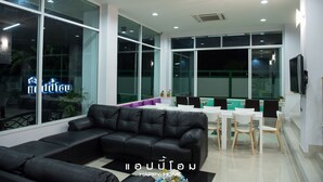 Uma sala de estar moderna com um sofá de couro preto, uma área de jantar com mesa e cadeiras brancas, e janelas amplas com vista para o exterior.