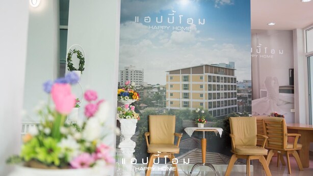 Um saguão de hotel com um grande cartaz exibindo a frase "HAPPY HOME" e texto tailandês, duas cadeiras de madeira, uma mesa pequena e uma planta em vaso com flores.