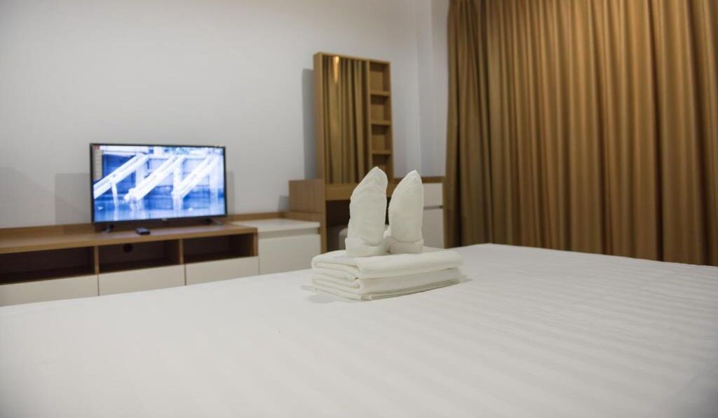 Quarto de hotel com cama bem arrumada, televisão sobre suporte de madeira e uma janela grande com cortinas.