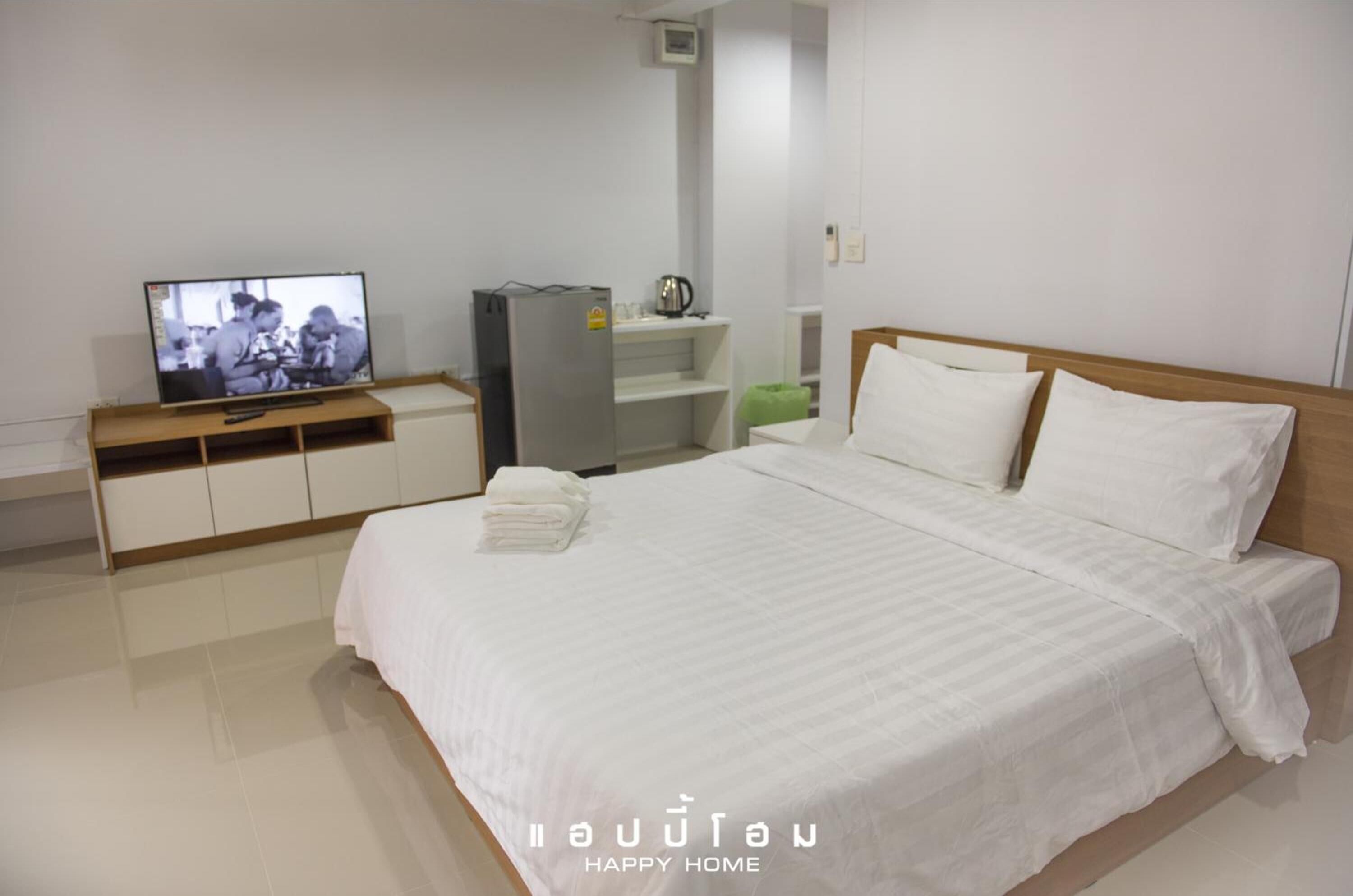 A hotel room with a bed, a television, a refrigerator, and a shelf with a kettle.