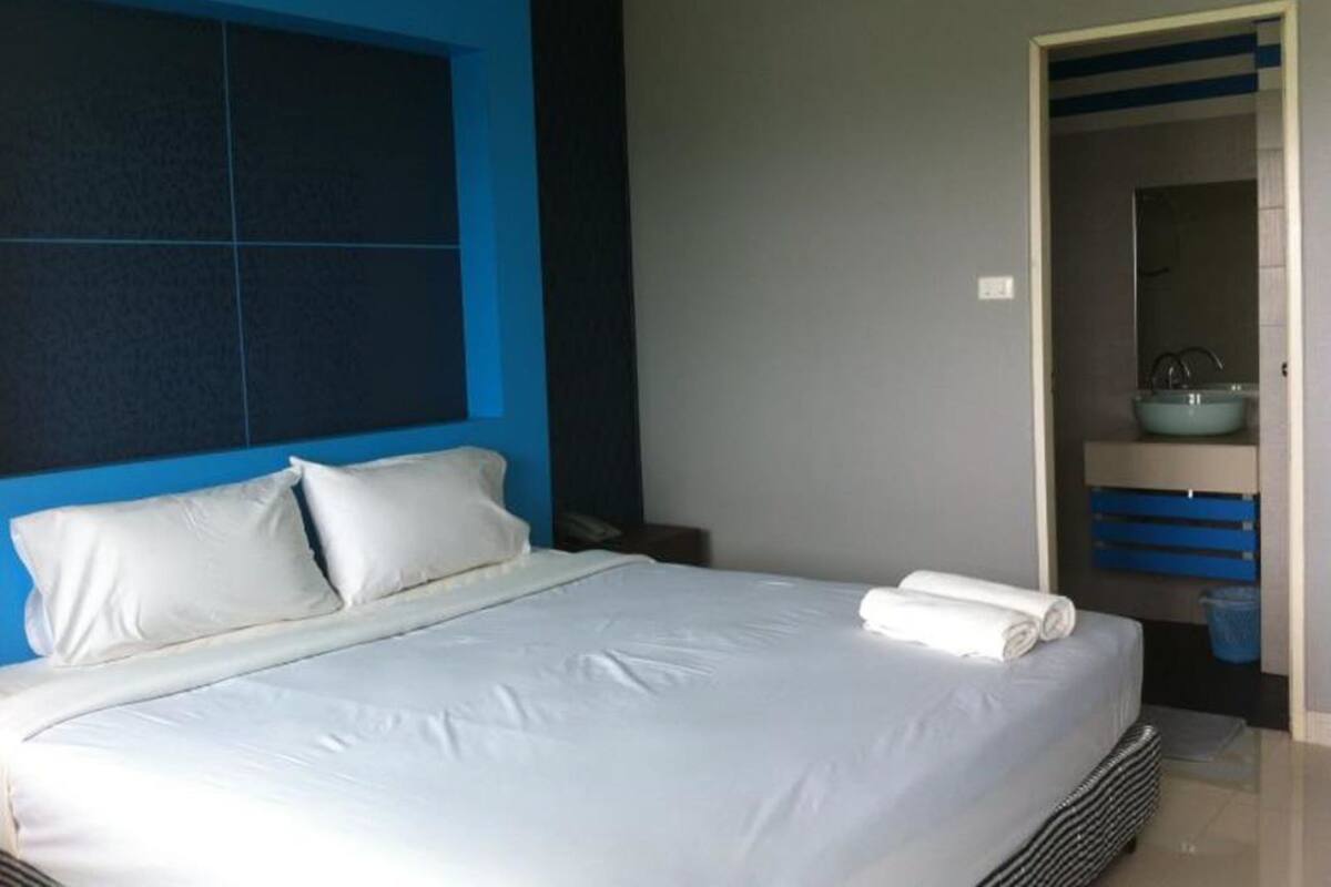 Ett modernt hotellrum med en stor säng, en blå accentvägg, ett litet badrum med handfat och spegel samt en snyggt bäddad säng med vita sängkläder.