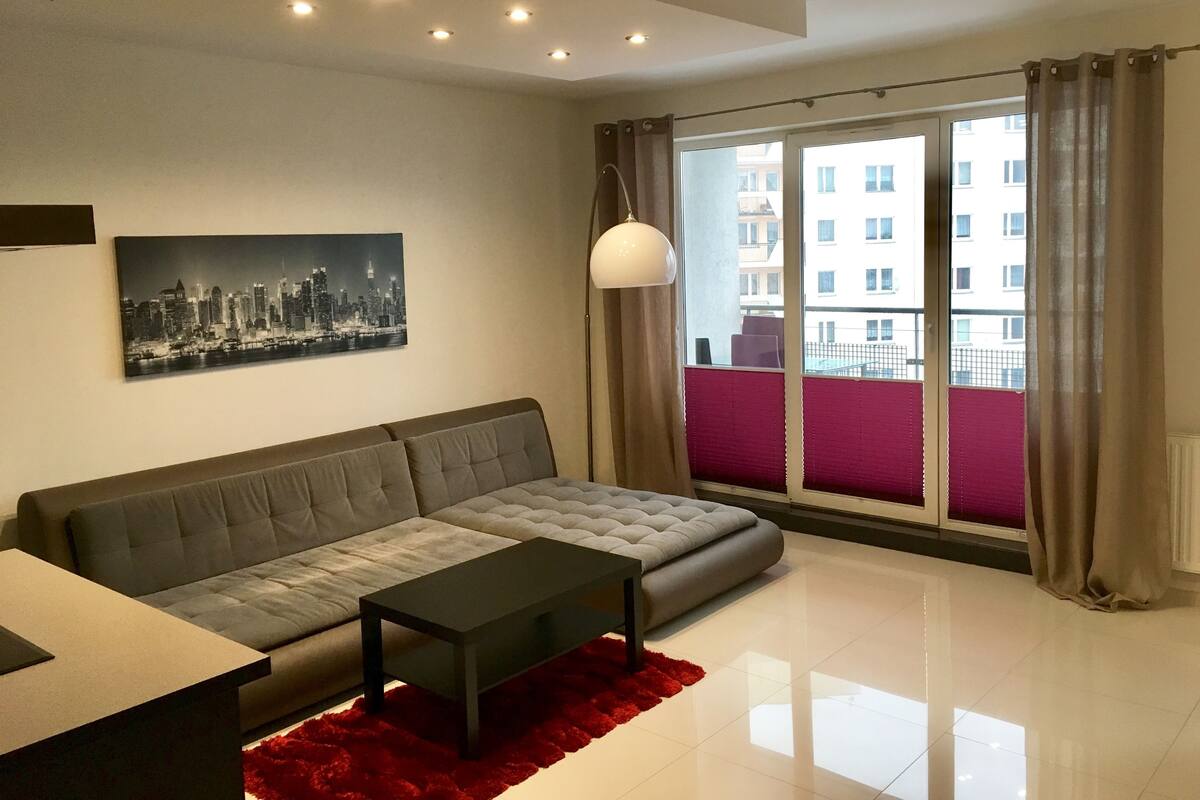 Una sala moderna con un sofá gris, una mesa de centro negra y un tapiz rojo. En la pared hay un cuadro de un paisaje urbano y un ventanal con cortinas.